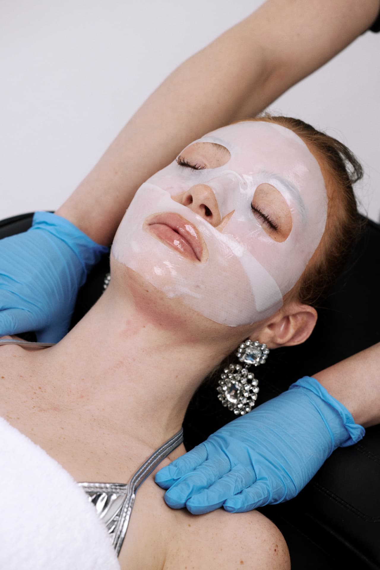 A client resting with a hydrating sheet mask on her face while a provider in blue gloves attends to her